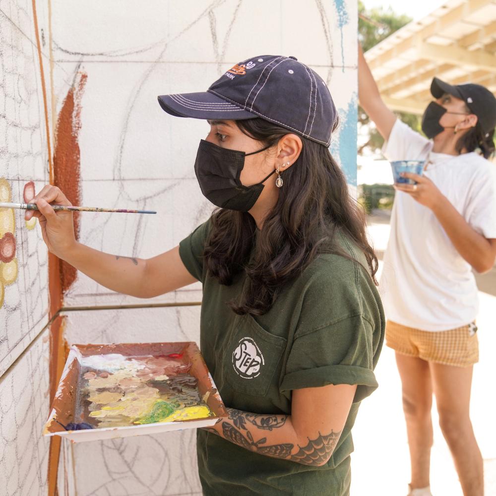 A student wearing a cap and facemask paints an outdoor mural