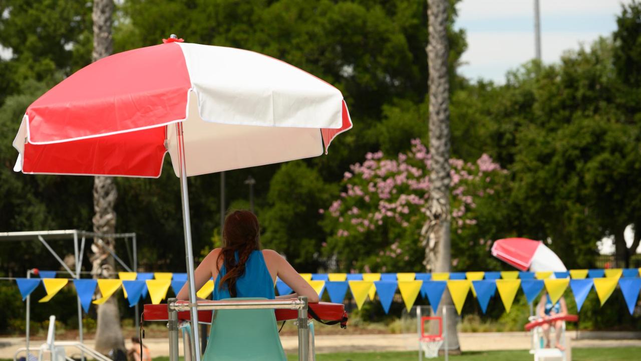 Lifeguard on high chair under red-and-white umbrella by outdoor pool, bunting flags