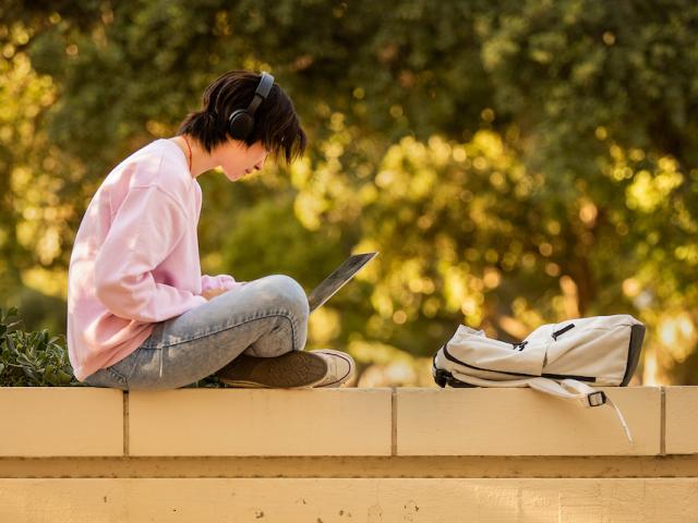Student with laptop sitting on an outdoor wall.