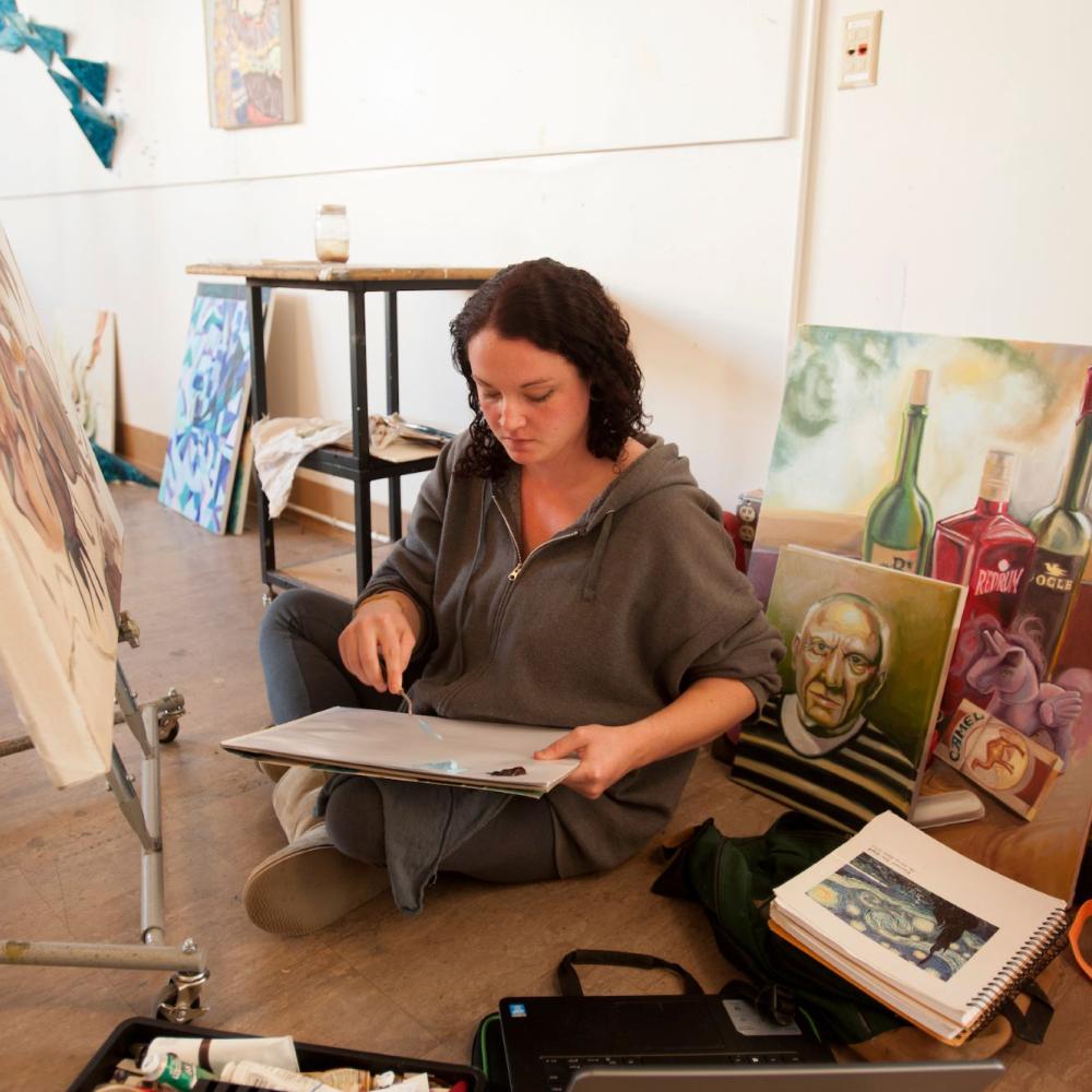 Woman sketching on floor in art studio, surrounded by canvases and supplies
