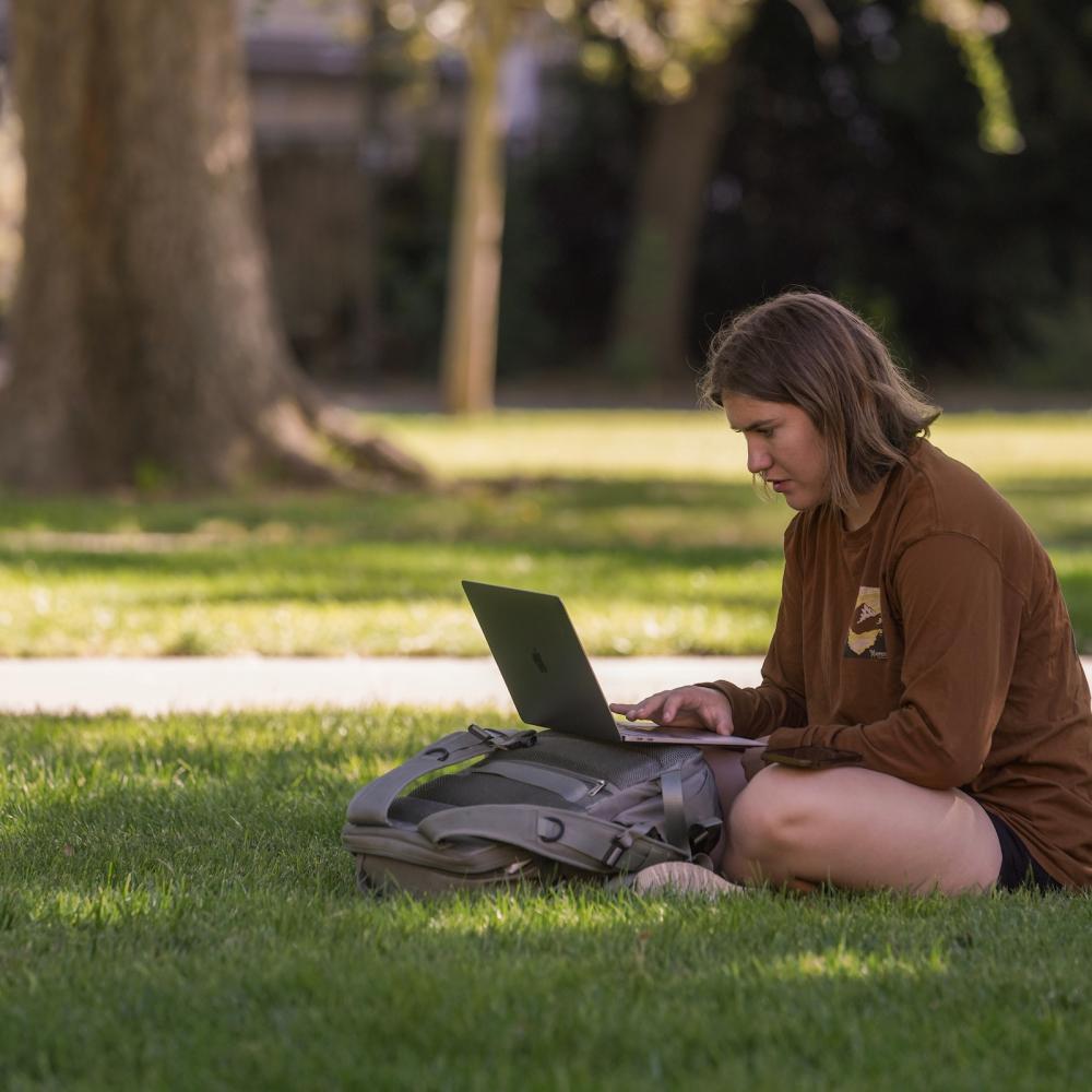 A student sitting on the grass with a laptop and backpack