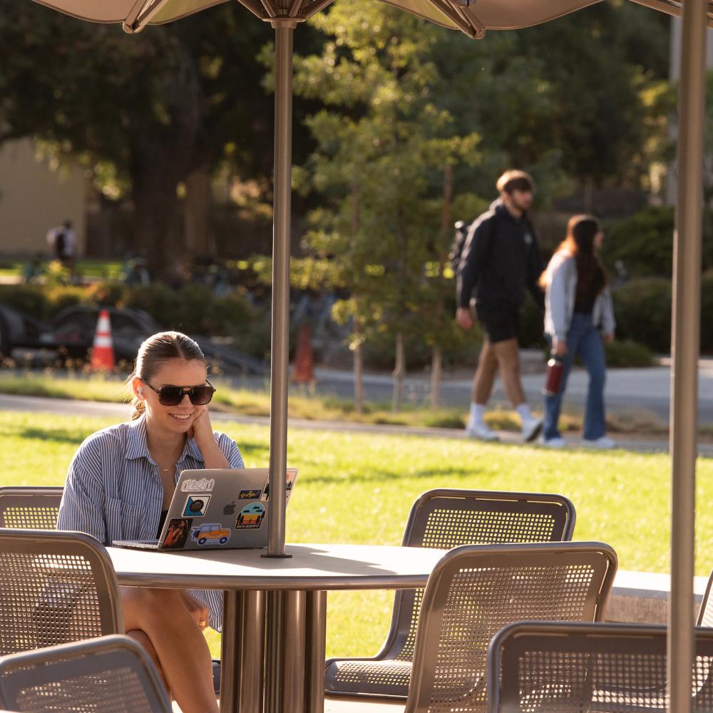 Student wearing sunglasses sitting outside at a table with a laptop