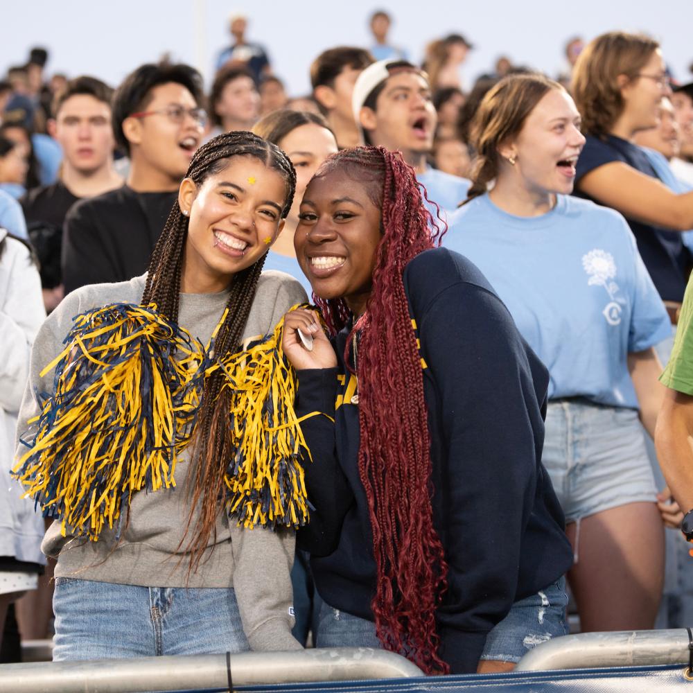 Two young women smile together at a lively event, surrounded by a cheering crowd.