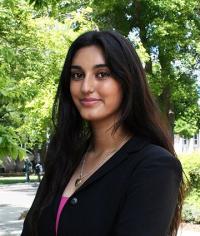 Headshot of a smiling young woman in a black blazer and pink top, leafy outdoor background