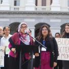 Aridin speaks at a podium in a hot pink outfit in front of a government building, surrounded by colleagues with signs that read “Education shouldn’t be a privilege” and “Budget better have my money.”