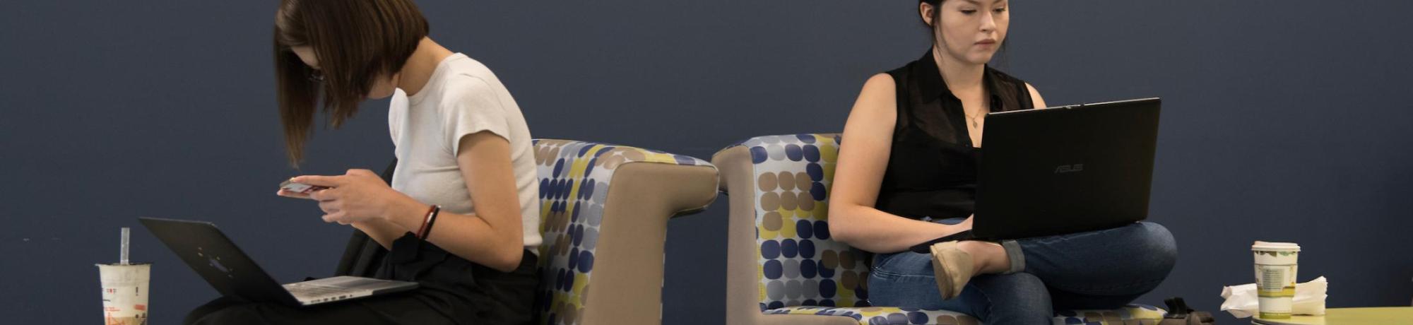 Two women back-to-back on patterned lounge seating, each using a laptop, drinks at sides.