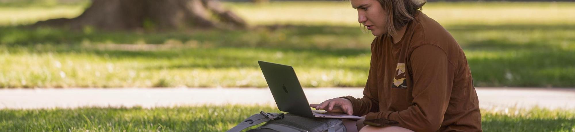 A student sitting on the grass with a laptop and backpack