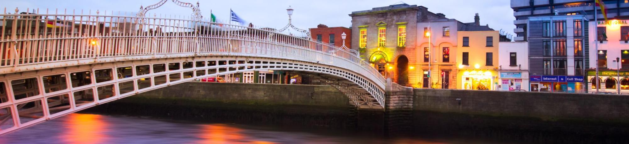 Ornate pedestrian bridge over river at dusk, colorful riverside buildings and reflections